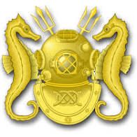 A gold embroidered or gold metal pin with two upright sea horses facing a diving helmet, and two tridents projecting upward and canted outward from the diving helmet's cover. A double carrick bend superimposed on the breast plate.